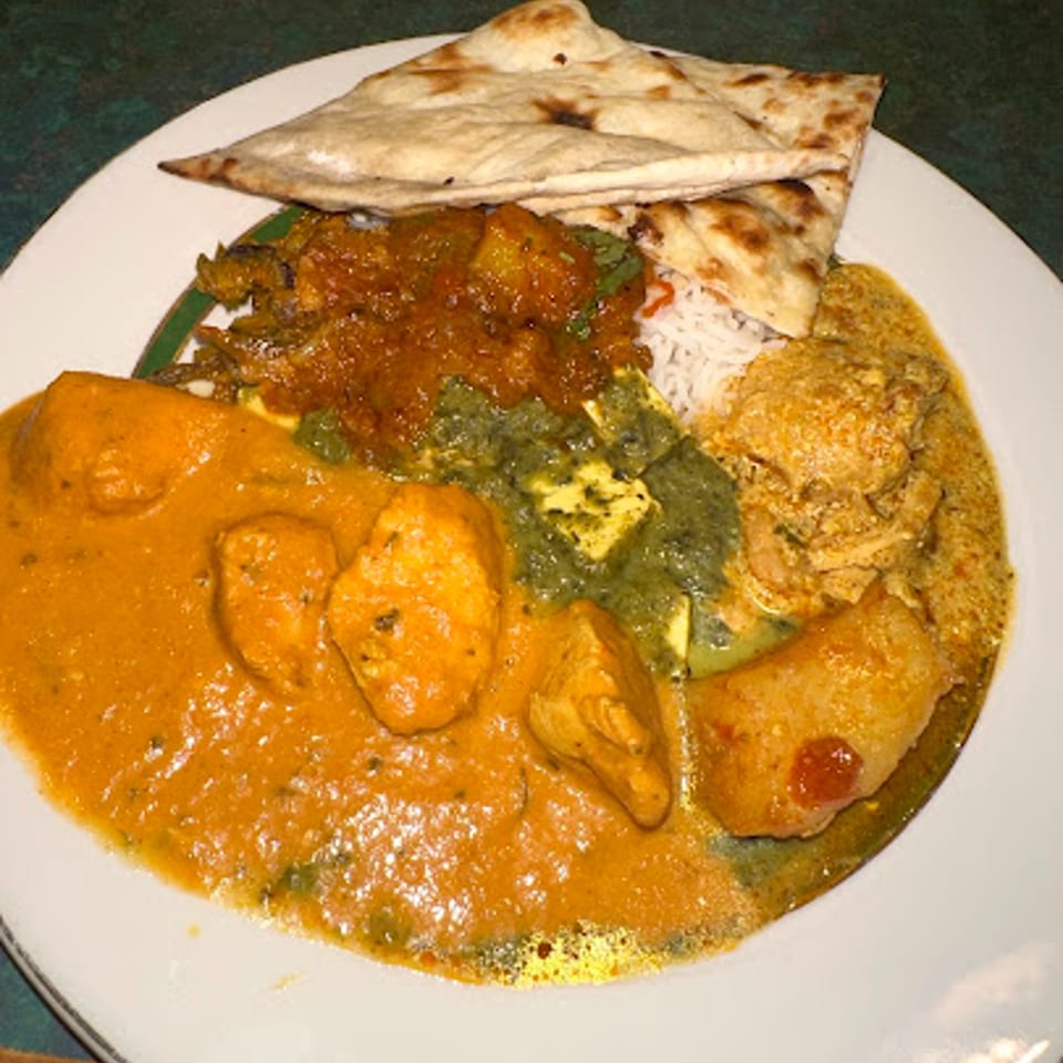 A white plate filled with various Indian dishes, including chicken curry, palak paneer, rice, and naan.