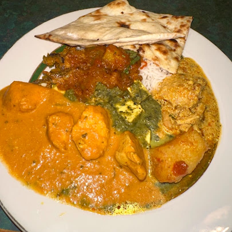 A white plate filled with various Indian dishes, including chicken curry, palak paneer, rice, and naan.