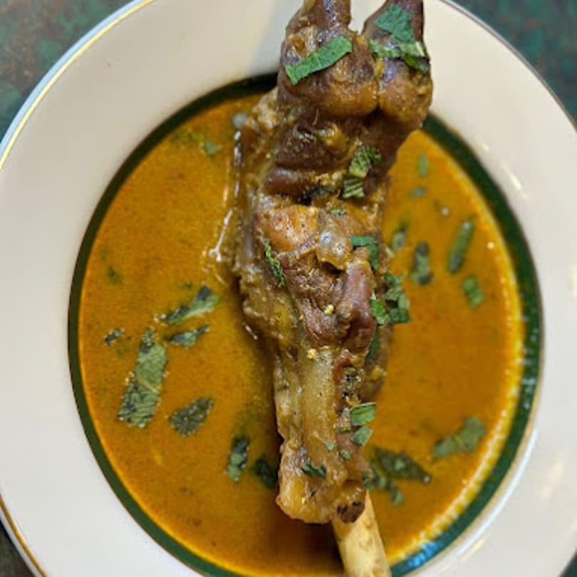 A close-up of a lamb shank in a rich, orange curry sauce, garnished with fresh herbs.