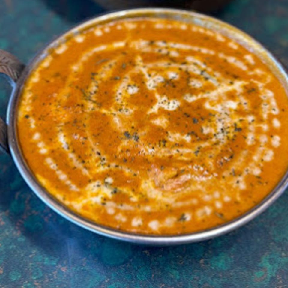 A close-up of a vibrant orange Indian curry, garnished with cream swirls and dark spices in a metal bowl.
