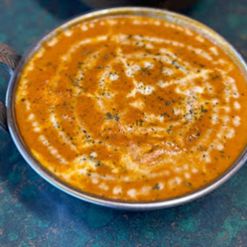 A close-up of a vibrant orange Indian curry, garnished with cream swirls and dark spices in a metal bowl.
